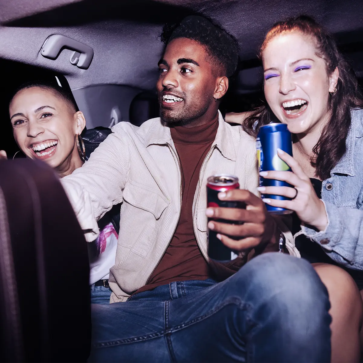 Group of friends laughing with drinks in the back seat of a car at night.