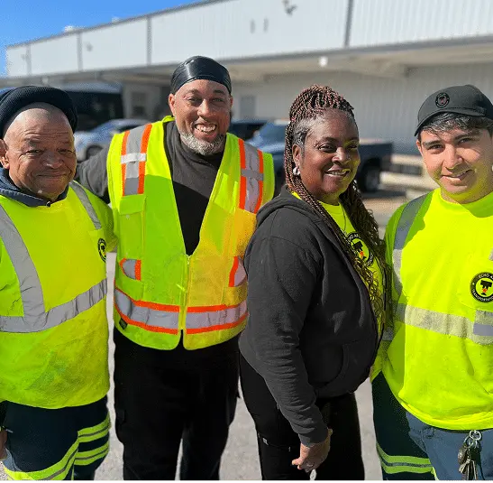 Team of four employees in high-visibility safety gear smiling outside a warehouse.