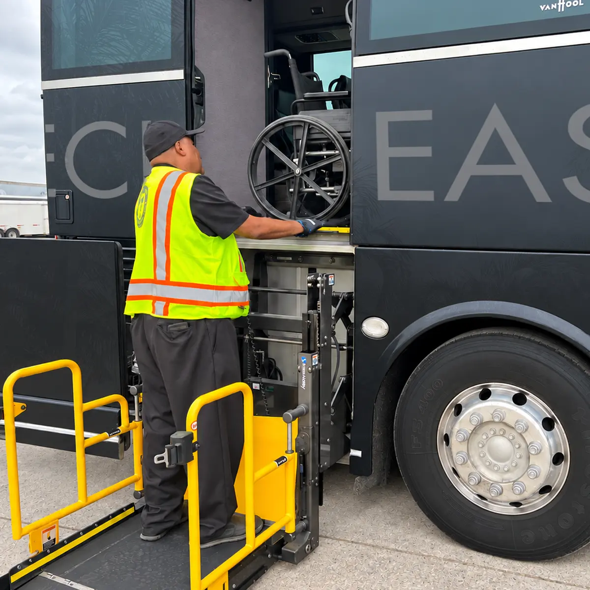Attendant in a safety vest loading an empty wheelchair onto a bus's wheelchair lift.